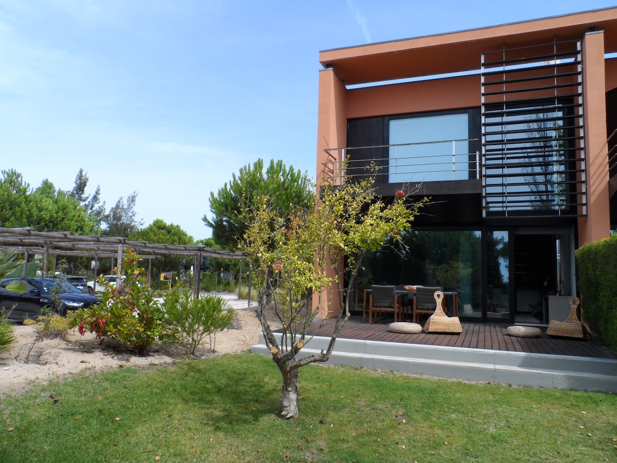The exterior of a modern town house is depicted, featuring a mix of sleek lines and natural elements. A small tree is visible in the foreground, with a grass area leading up to the house. Large windows provide a view into the interior, enhancing the connection with nature.