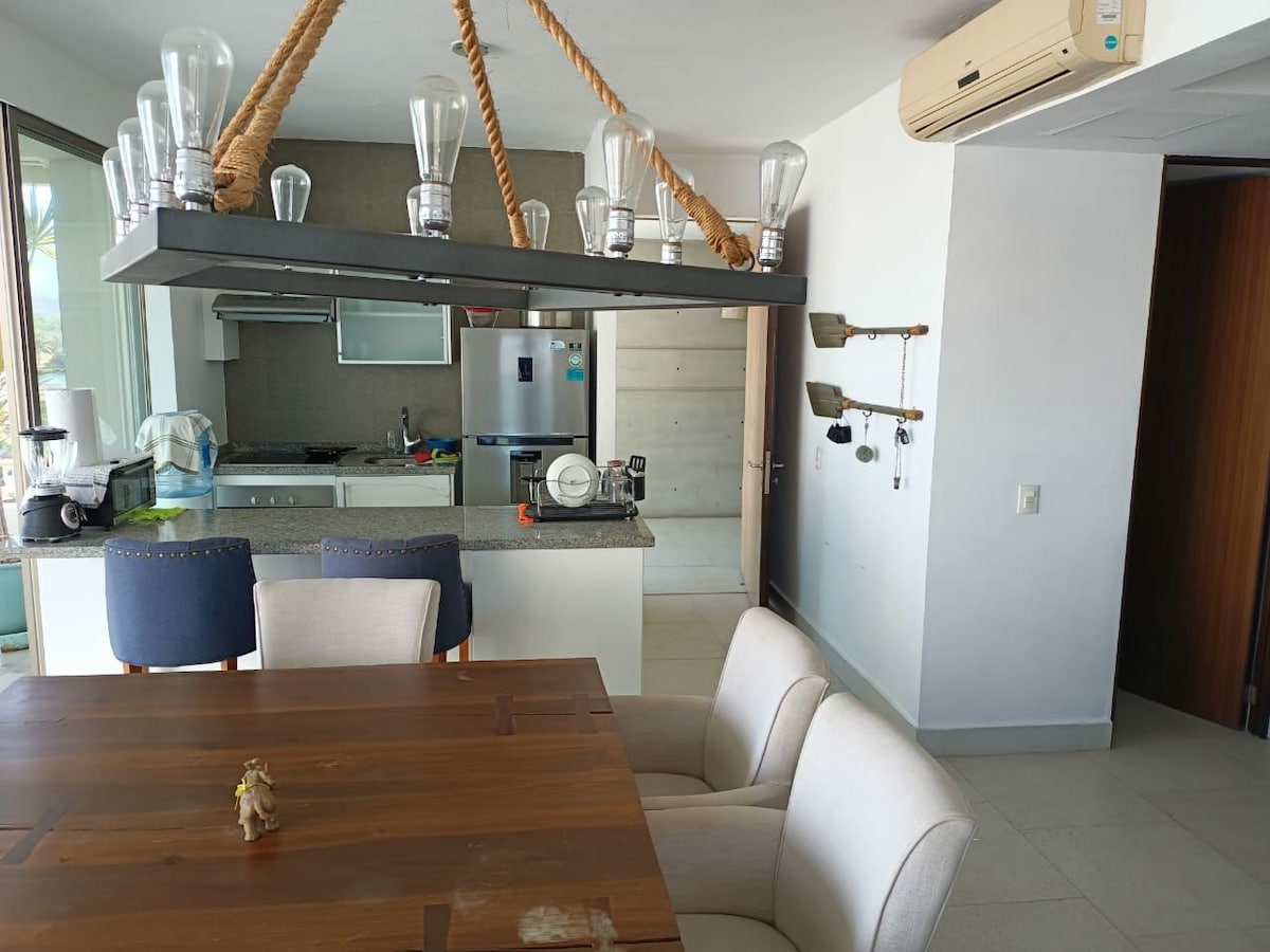 A modern dining area features a rustic wooden table accompanied by two upholstered chairs. Above, a unique light fixture made of glass bulbs is suspended from a rope. The kitchen area is visible in the background, showcasing sleek cabinetry and stainless-steel appliances.