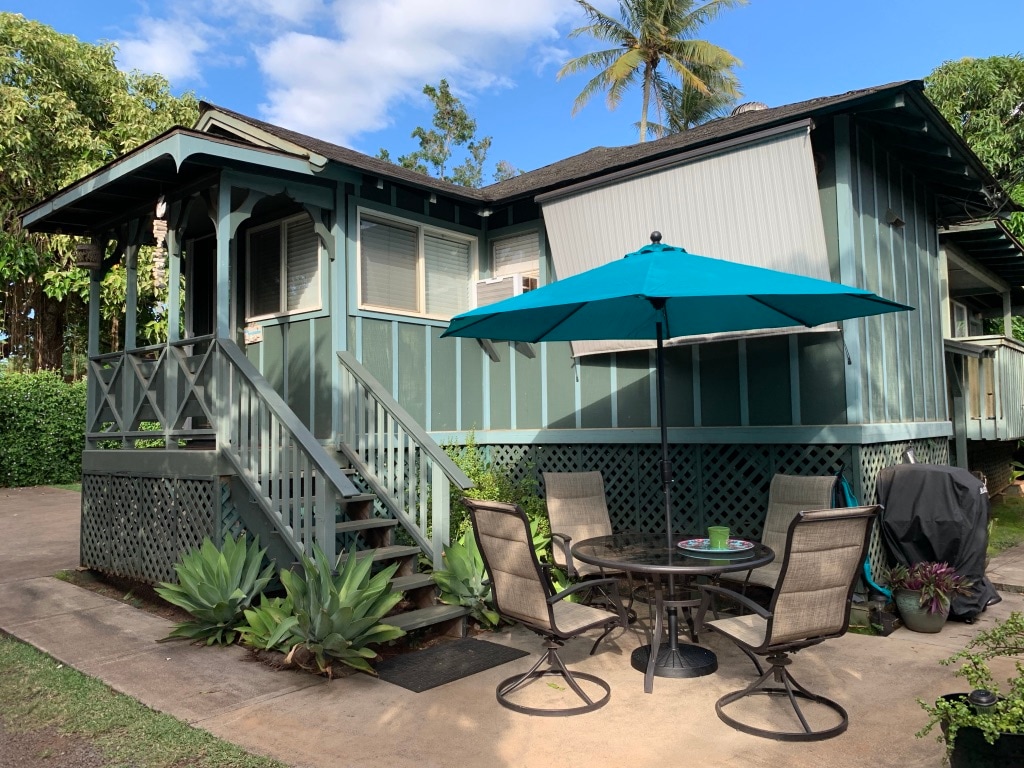 The exterior of the house is presented, featuring a green façade with decorative lattice and a set of stairs leading to the entrance. A round table with four chairs is situated outside, shaded by a teal umbrella, complemented by surrounding tropical plants.