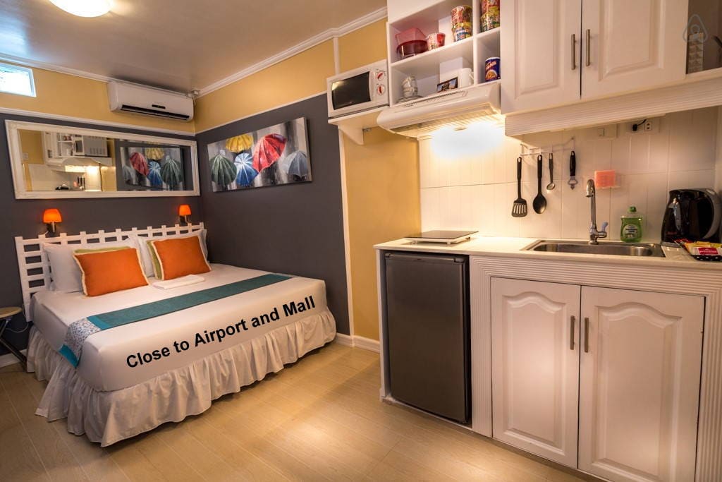 A compact room features a clean bed with colorful accent pillows. A well-equipped kitchen area includes a microwave, kettle, and refrigerator. A mirror reflects vibrant artwork on the wall, and light from a window brightens the space, creating a functional atmosphere.