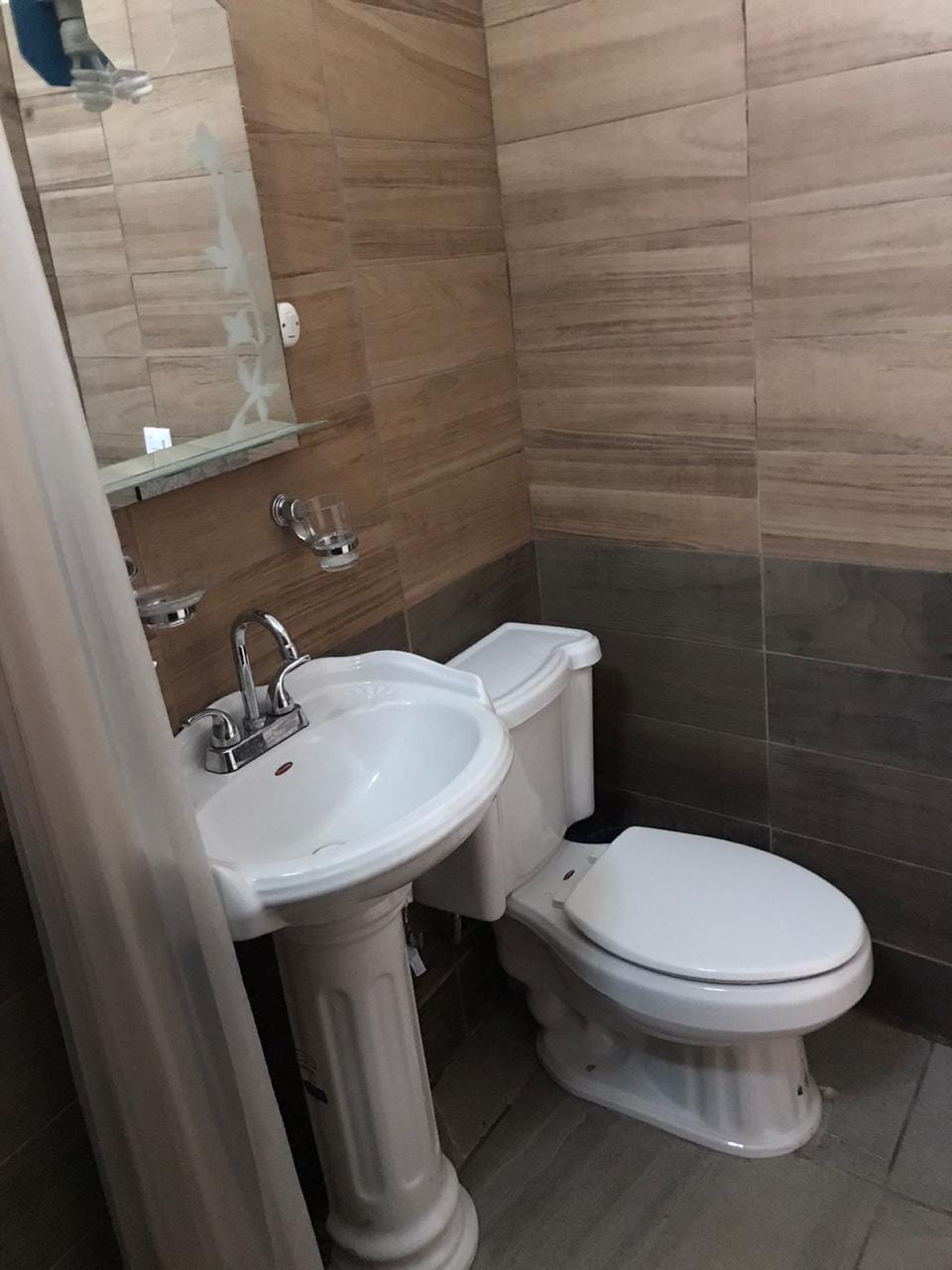 A compact bathroom features a white sink and a modern toilet, positioned against a backdrop of textured wooden tiles. A mirror is mounted above the sink, reflecting the neutral tones of the space. The floor is tiled in a coordinating shade, enhancing the overall design.