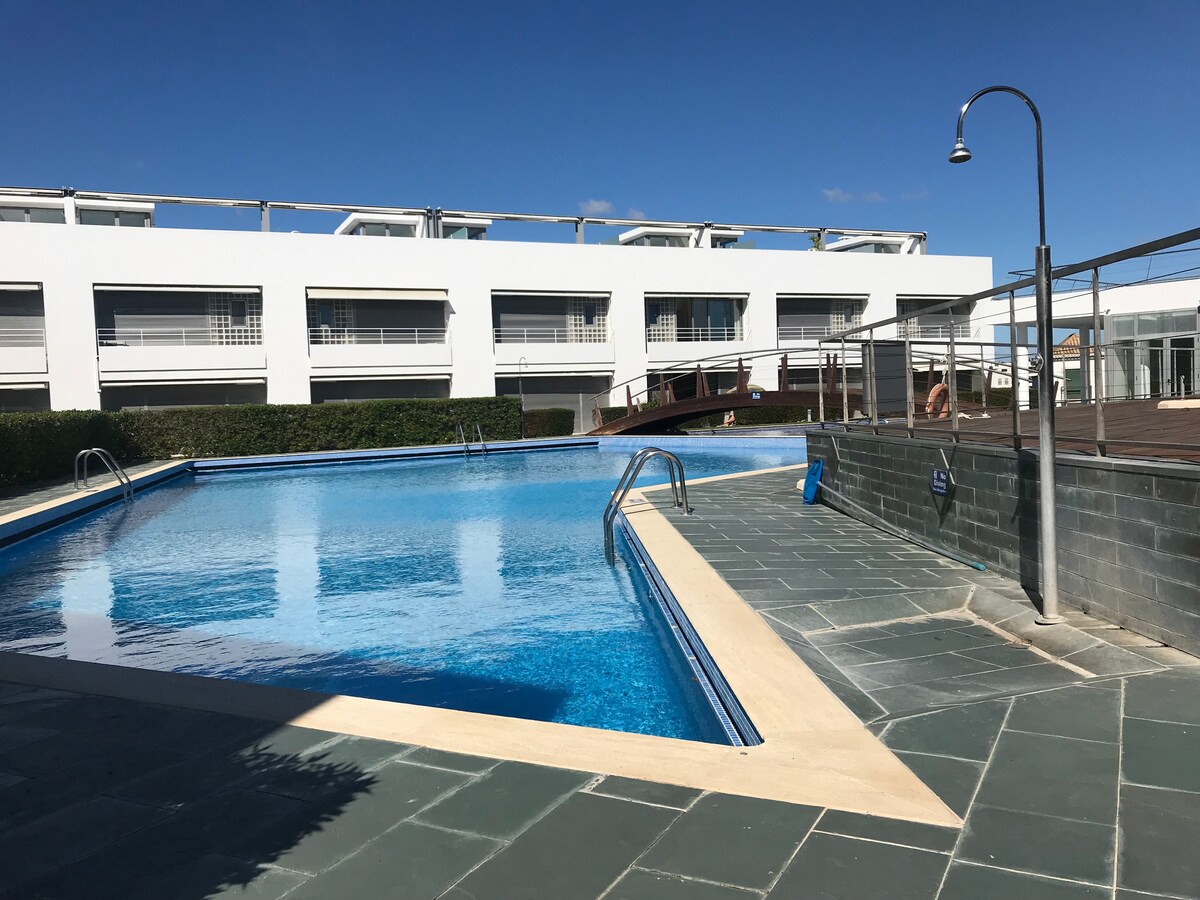 A swimming pool is viewed, featuring a clear blue surface reflecting the sky. The pool's shallow entrance is lined with steps. Surrounding it are neatly maintained green hedges and a wooden deck area, providing a sense of openness and tranquility.