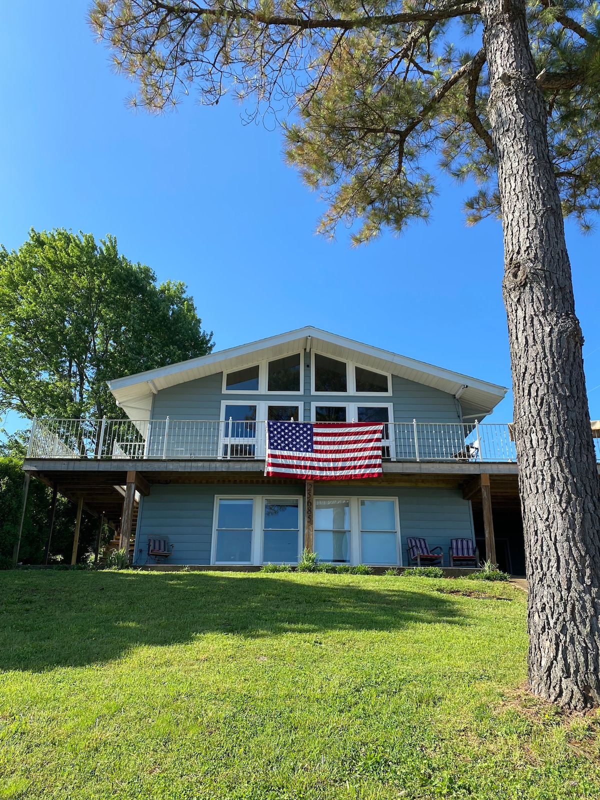 A two-story house features a large deck with a prominent American flag displayed. Large windows illuminate the front, while green grass and trees surround the property, creating a serene outdoor environment. The home showcases a contemporary design with a triangular roof.