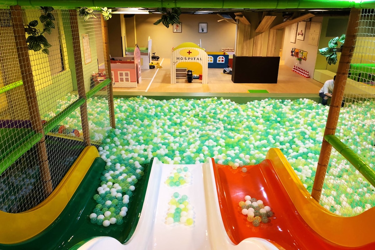 A vibrant play area is displayed, featuring two colorful slides in orange and white. The floor is covered with a sea of soft, multicolored balls in green and white. Various play structures can be seen in the background, enhancing the playful environment.