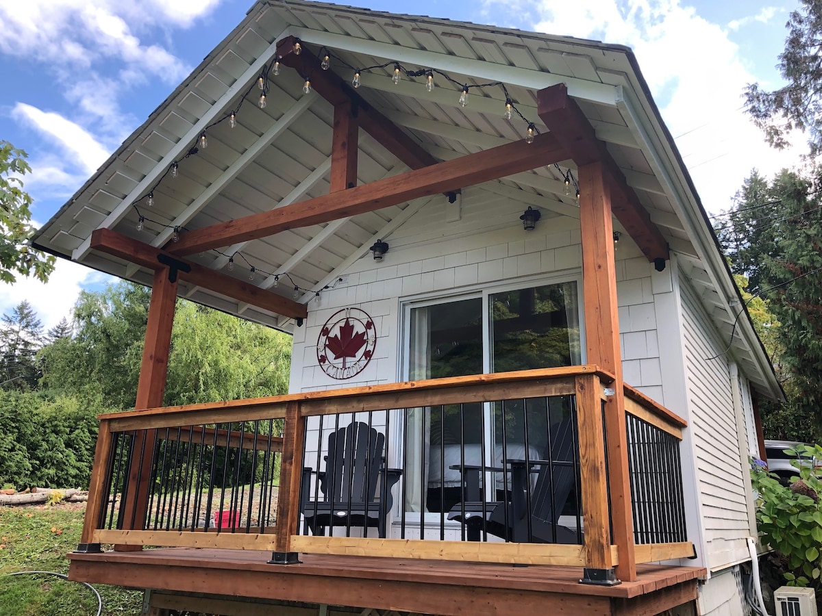 A charming exterior of the cabana displays a large covered porch supported by wooden beams. Black outdoor chairs are positioned on the deck, inviting relaxation. A decorative emblem is mounted on the wall, and large windows offer a view into the inviting interior space.