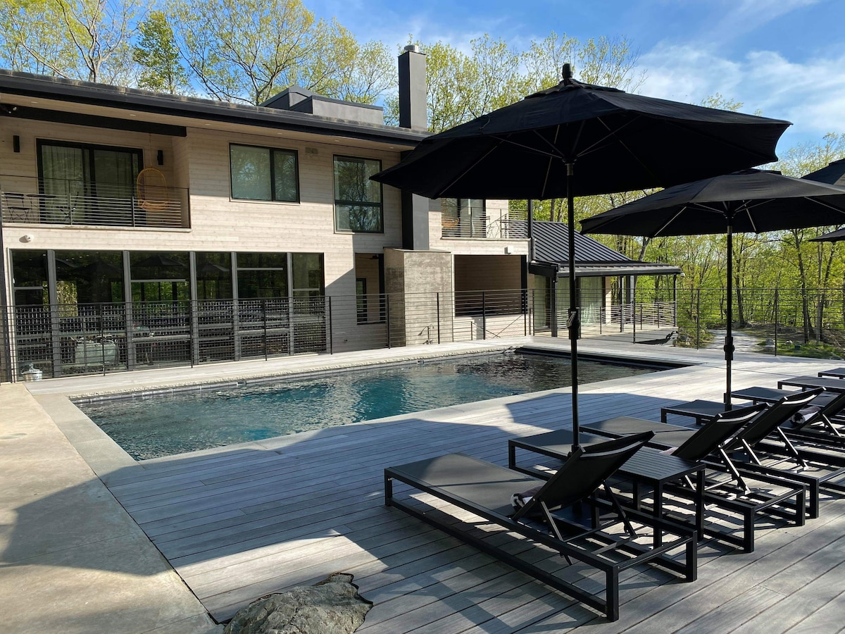 A spacious outdoor area features a modern pool surrounded by wooden decking. Lounge chairs are positioned under large umbrellas, providing shade. The two-story home is visible in the background, showcasing large windows that enhance natural light.