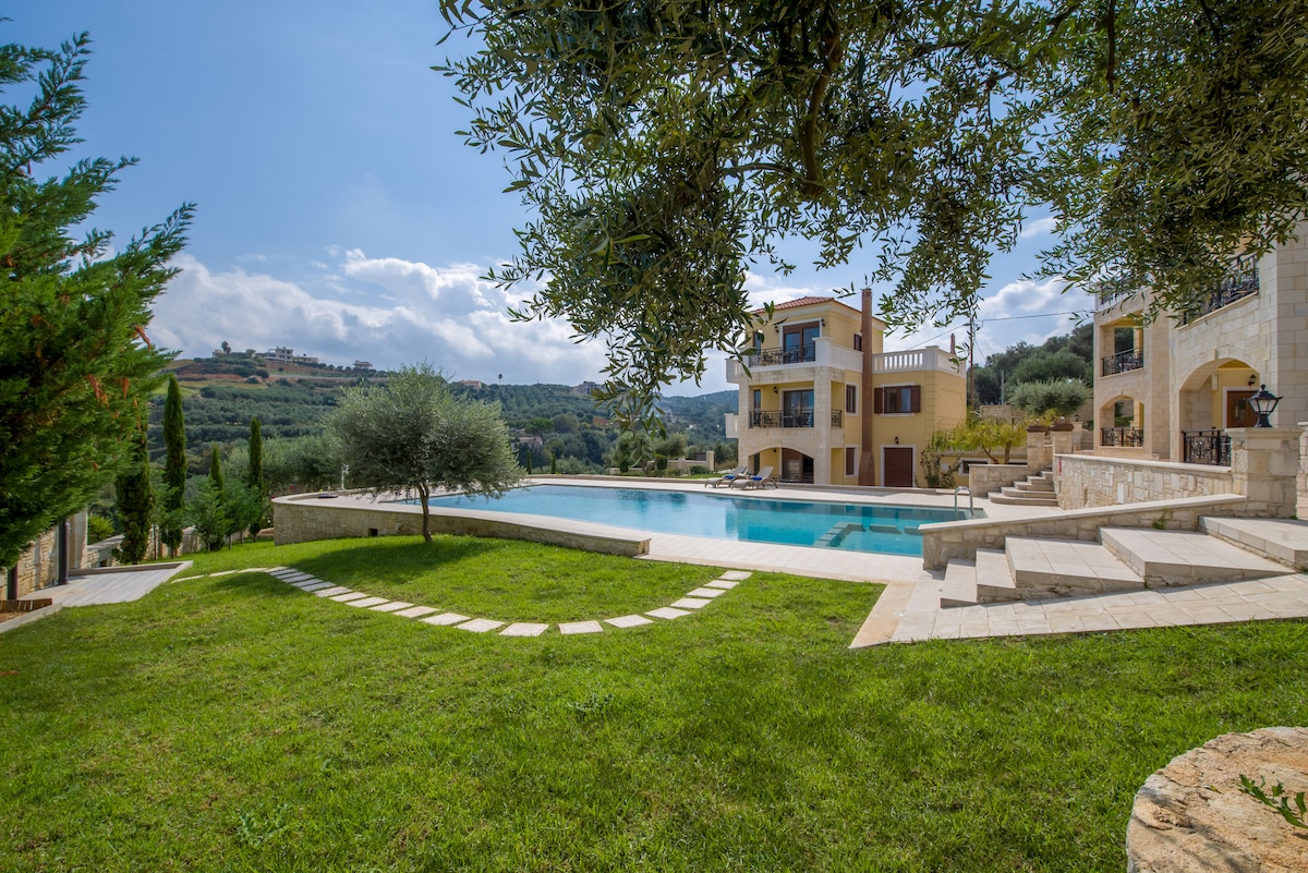 An expansive outdoor area is depicted, featuring a large pool surrounded by a landscaped garden. The villa stands in the background, with multiple floors visible. Neatly cut grass leads to stone pathways and mature trees provide shade, enhancing the serene atmosphere.