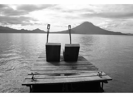 A wooden dock extends over the calm waters of Lake Atitlan, featuring two rustic chairs positioned side by side. The serene lake reflects the soft clouds above, with the silhouette of a volcano in the background, creating a moment of tranquility.