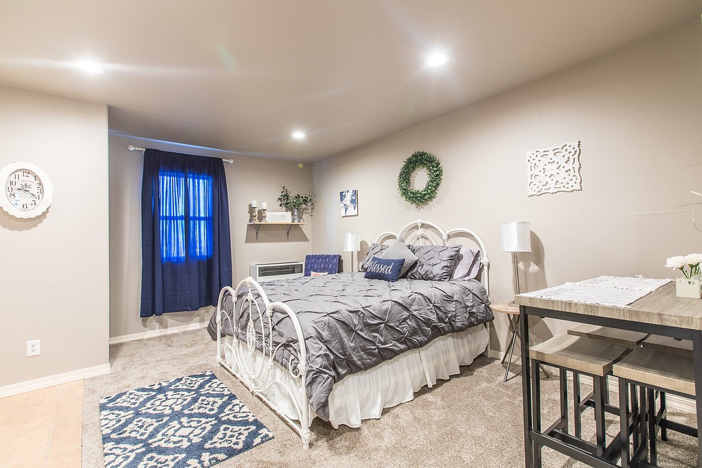 A spacious sleeping area features a queen bed with light bedding and decorative pillows. Nearby, a small dining table for two is positioned on a textured rug. A window with dark curtains allows natural light, complemented by wall decorations and greenery on the shelf.