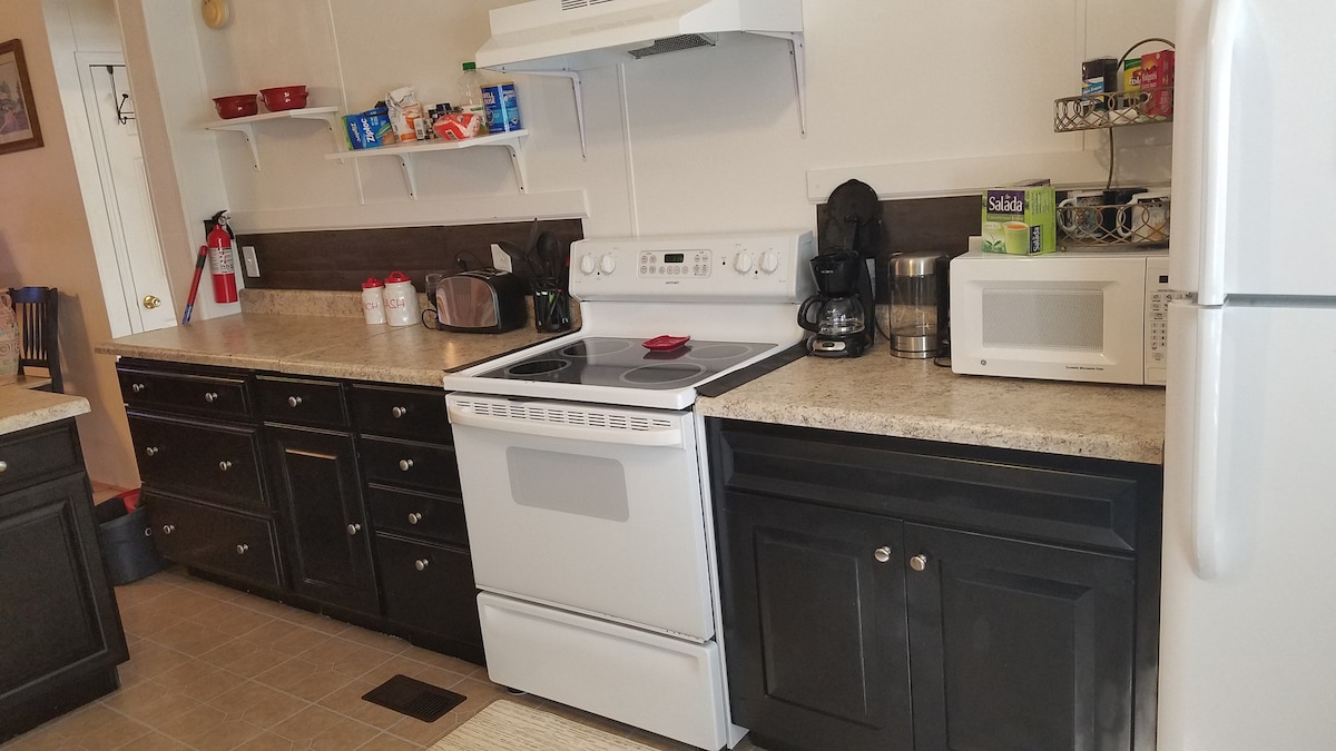 The kitchen features a mix of black cabinetry and light-colored countertops. A stove with an oven is positioned underneath a vent hood. Additional appliances include a microwave, coffee maker, and refrigerator, all arranged neatly for convenience. Open shelving holds utensils and storage containers.