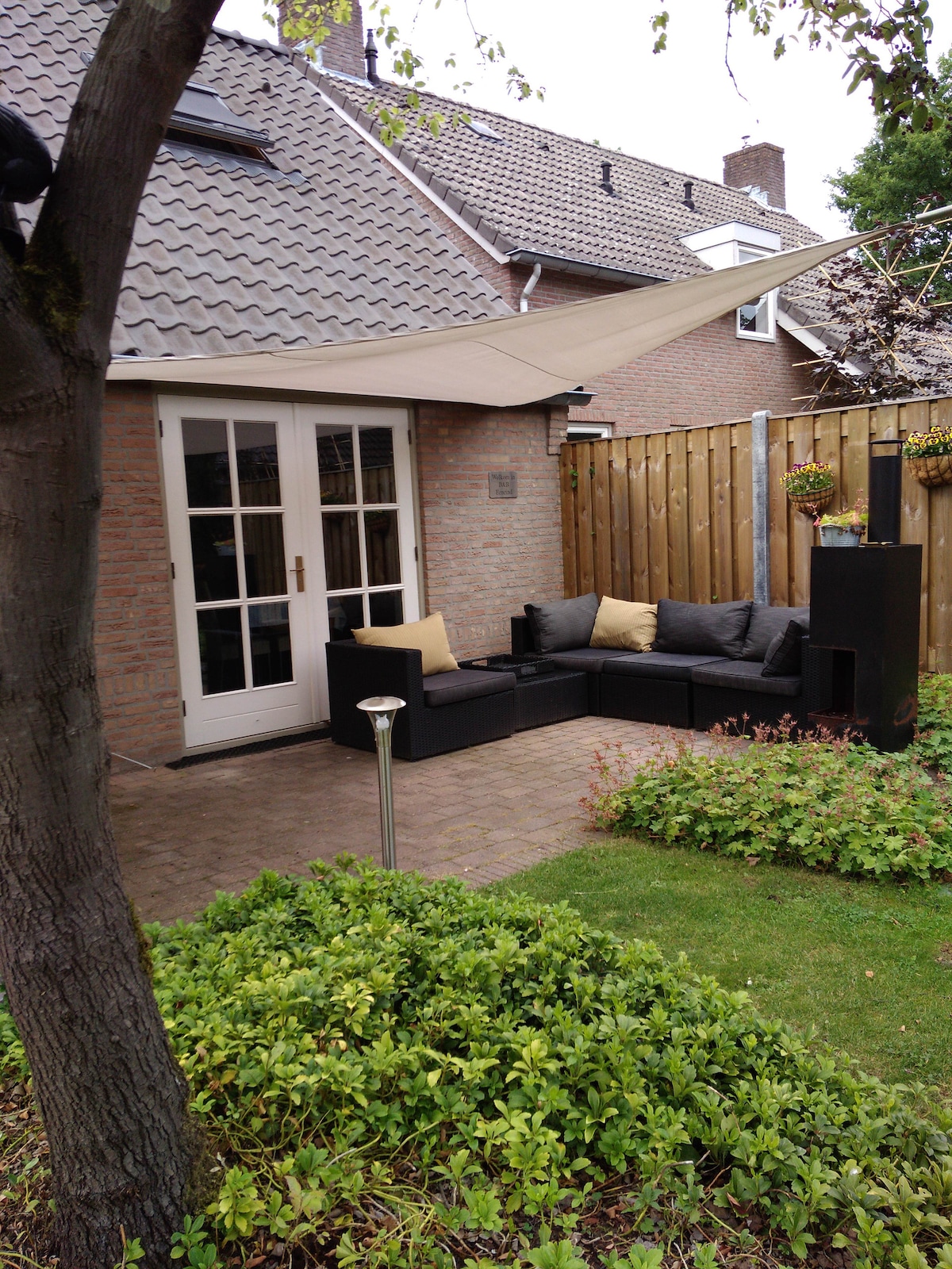 An outdoor seating area is visible, featuring a sectional sofa with plush cushions. A small table is placed nearby, with a decorative light fixture illuminating the space. A shaded canopy provides cover, surrounded by a well-maintained grassy area and wooden fencing for privacy.