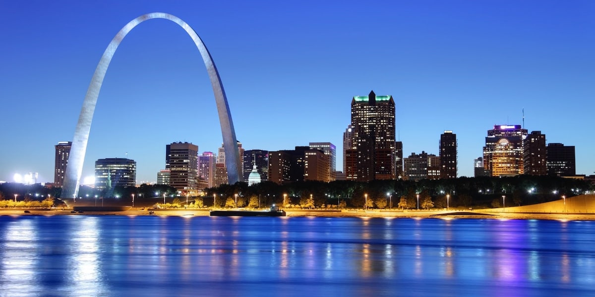 A nighttime view of the St. Louis skyline is captured, featuring the iconic Gateway Arch illuminated against a deep blue sky. The shimmering water reflects the colorful lights from the buildings, creating a vibrant urban scene.