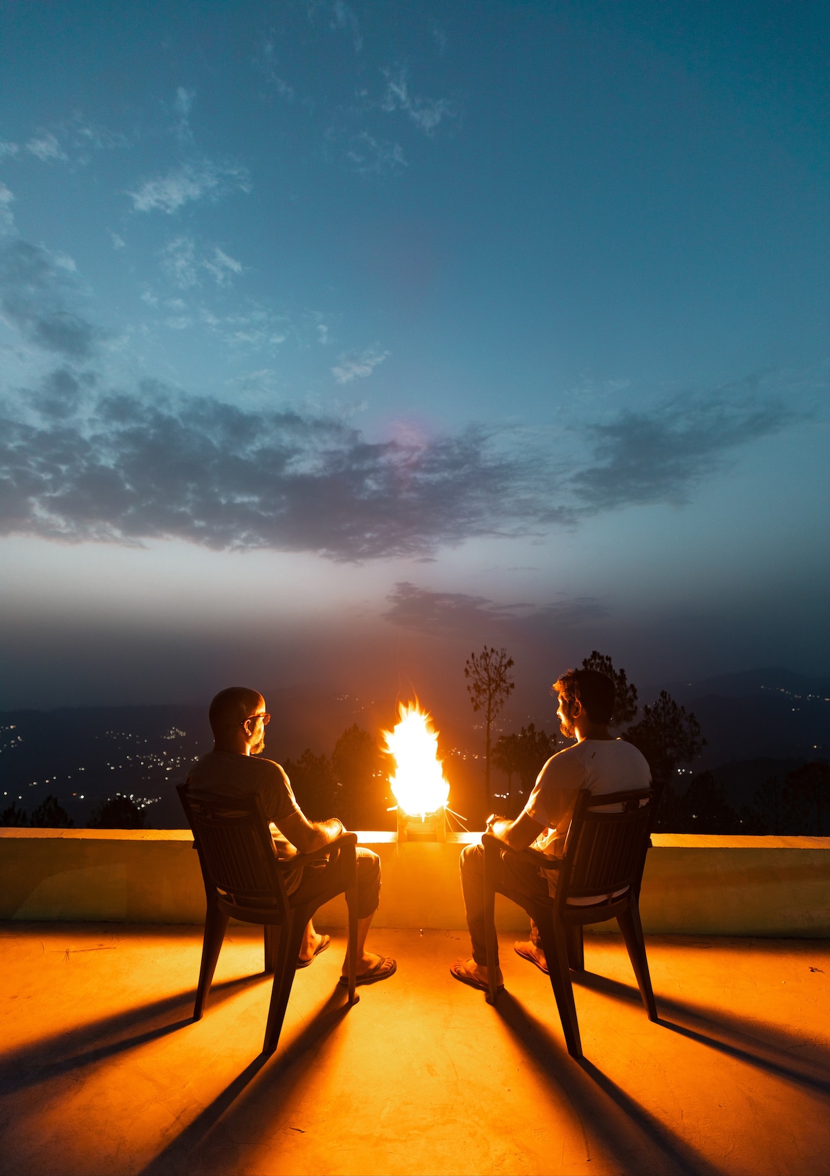 Two individuals sit on black chairs beside a fire pit on a terrace, with the evening sky transitioning from blue to dusk. Silhouetted against the horizon, the gentle flames provide warm illumination, creating a serene atmosphere that offers views of distant lights and mountains.