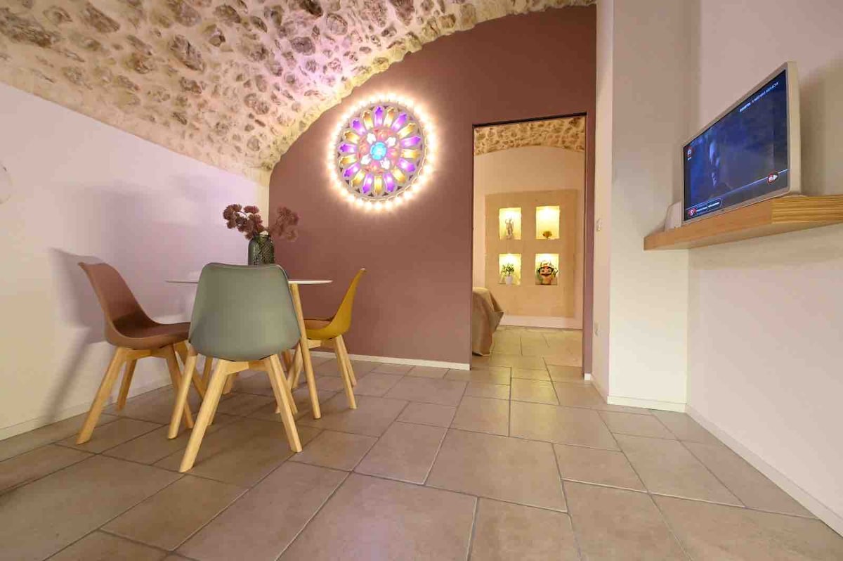 A dining area is featured with a round table and four chairs in varying colors. A decorative archway leads to a warmly lit bedroom, while a simple wall-mounted television is positioned above a wooden shelf. The ceiling displays exposed stone elements.