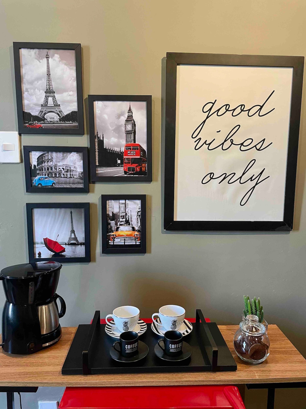 A decorative wall features a collection of black and white photographs of iconic landmarks, including the Eiffel Tower and a London double-decker bus. Below, a wooden table holds a coffee maker and two coffee cups on a black tray, complemented by a small plant and stylish decor.