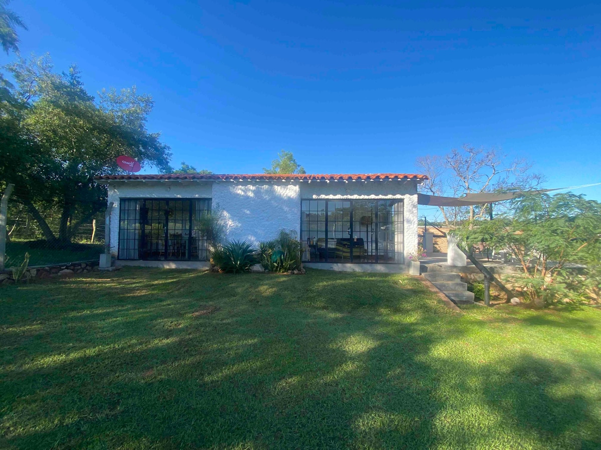 A single-story house features a rustic design with a tiled roof and large windows that provide ample natural light. Surrounding greenery enhances the outdoor space, and a well-maintained lawn offers a peaceful setting. A shaded area is visible, contributing to the serene atmosphere.