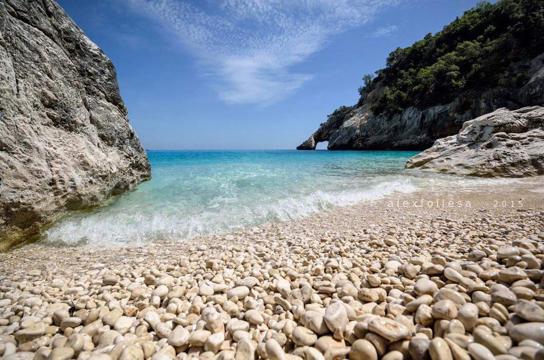 A serene beach scene showcases smooth, finely polished pebbles along the shoreline. The clear turquoise water gently laps at the beach, framed by rugged cliffs and vibrant greenery in the background under a bright blue sky.