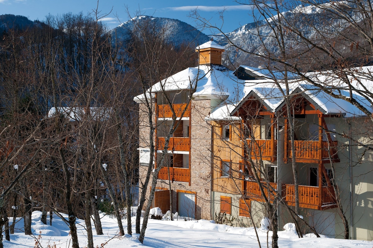 A charming multi-story building is surrounded by snowy trees and the quiet landscape. Wooden balconies are visible on the upper floors, highlighted against the snow-covered ground. The serene backdrop features distant mountains under a clear blue sky.