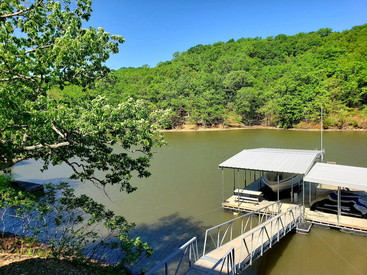 A serene view of a calm lake is framed by lush green trees, showcasing a wooden dock with a covered boat slip. The gentle slopes of the surrounding hills create a tranquil backdrop, reflecting the peacefulness of the natural setting.