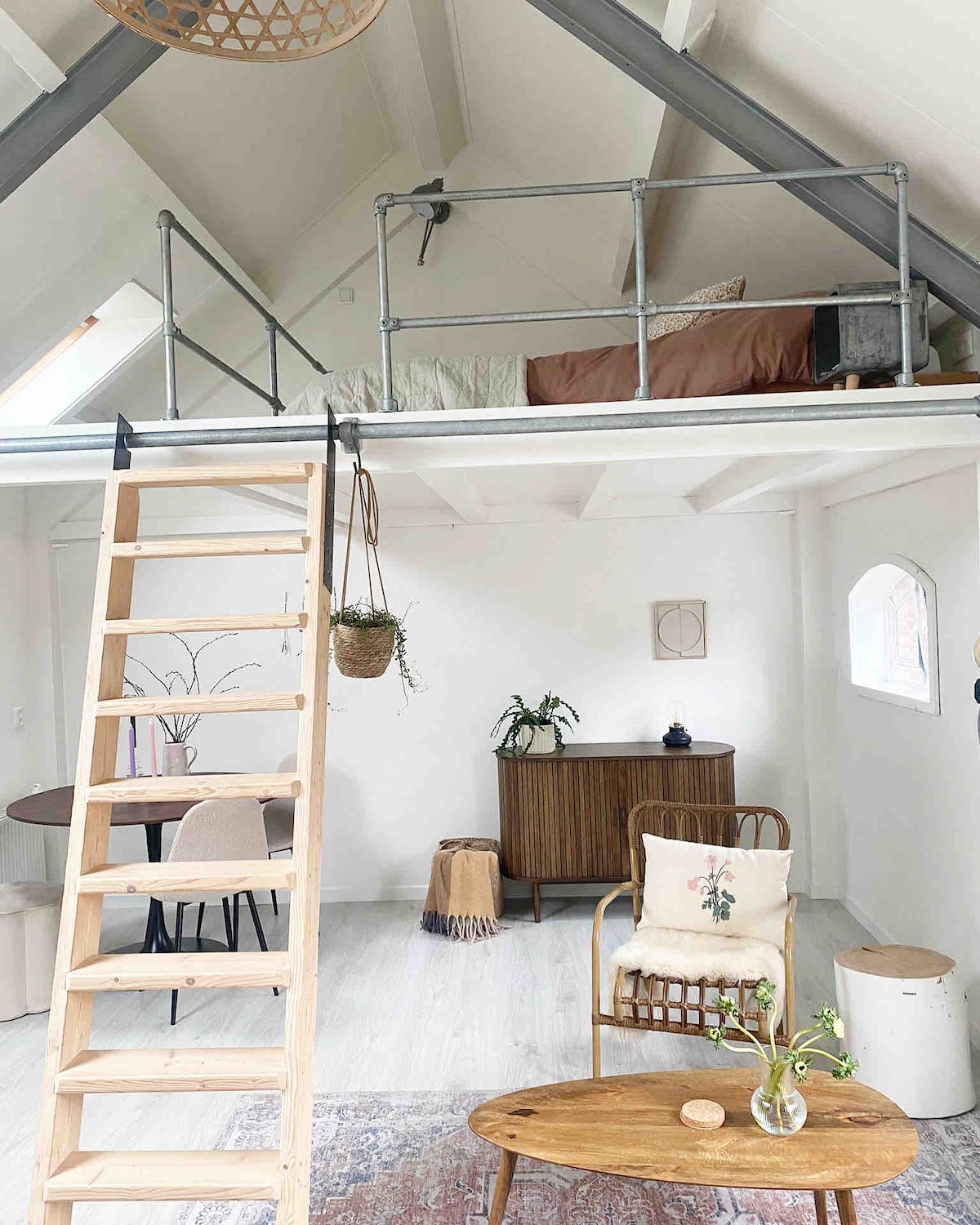 An open living space is highlighted by a cozy seating area and a wooden table with chairs. A staircase leads to a sleeping loft, enclosed by a simple railing. Natural light filters through a small window, illuminating the neutral decor and indoor plants.