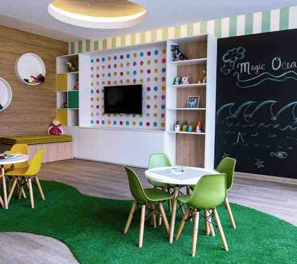 A colorful children's room features playful elements including a chalkboard wall with ocean-themed doodles. Green grass-like carpet enhances the lively atmosphere. Two tables with chairs in various shades surround the space, while a TV within a white cabinet adds to the functional design.