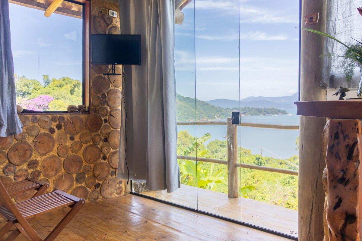 A cozy interior features a combination of wooden textures and stone walls, with large glass doors providing a clear view of the surrounding landscape. A television is mounted on the wall, and a folding chair is placed nearby, enhancing the welcoming atmosphere.