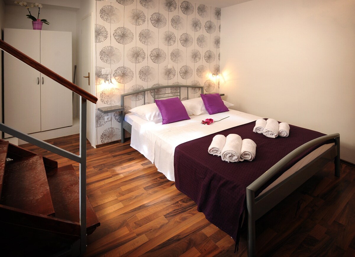 A sleeping area features a spacious bed dressed in white linens and accented by purple pillows. Fresh towels are neatly arranged at the foot of the bed. The room includes a staircase leading to an upper level, with floral wallpaper complementing the warm wood flooring.