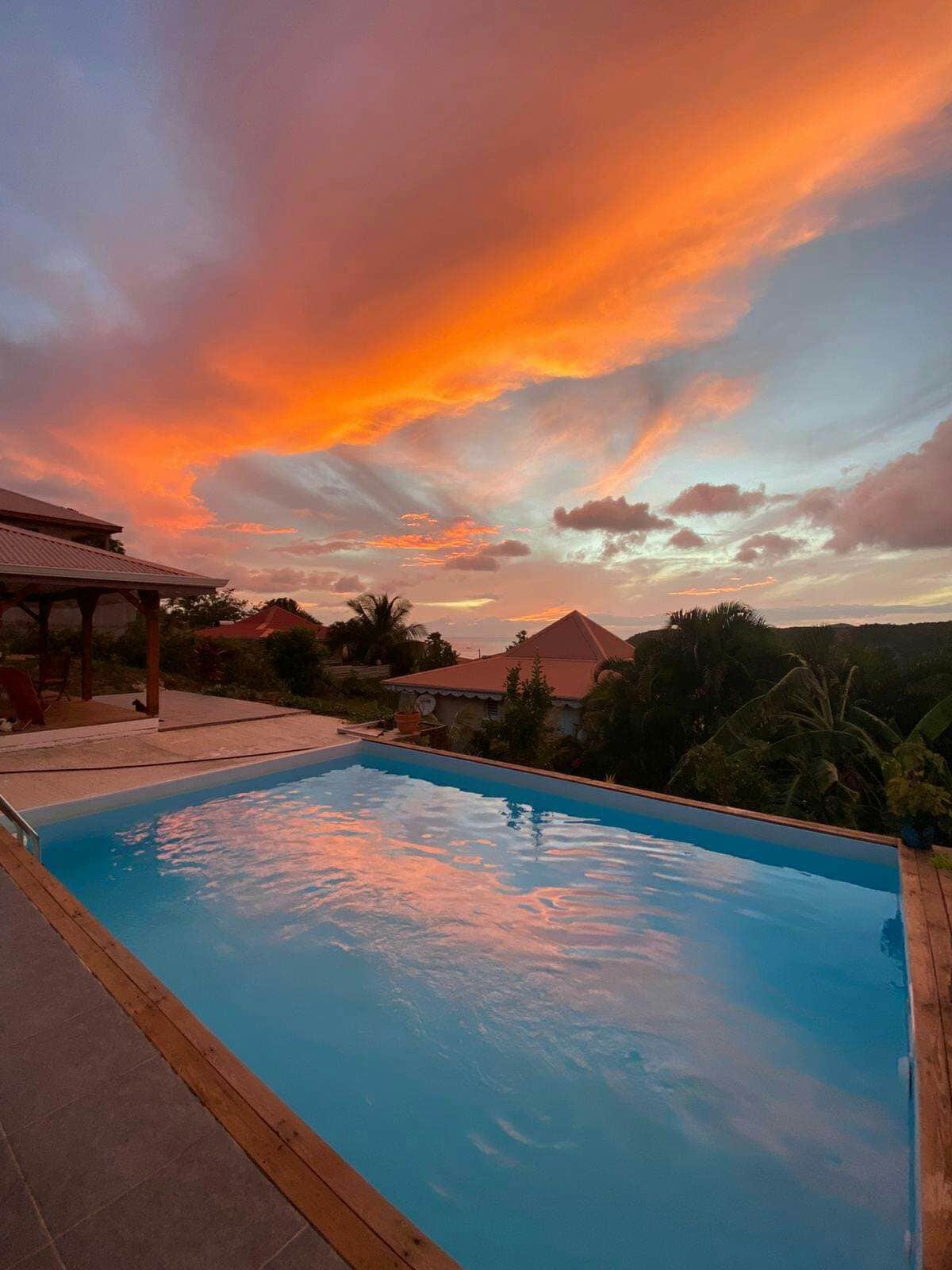 A serene swimming pool is framed by a wooden deck, reflecting the vibrant orange and pink hues of a sunset sky. Lush greenery surrounds the area, providing a natural backdrop to the tranquil scene.