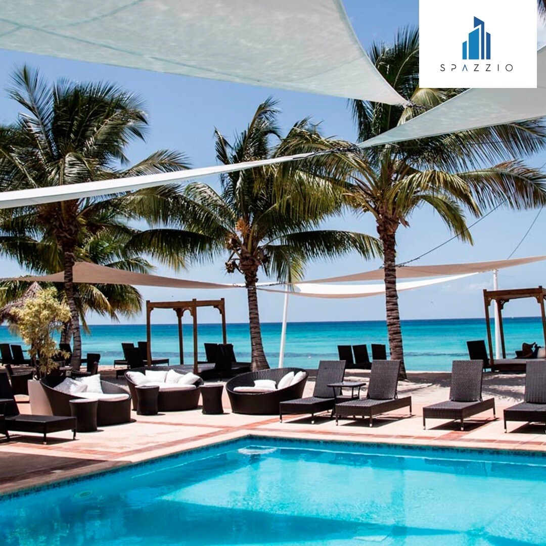 A sparkling pool is surrounded by lounge chairs and shaded areas under large canopies. Palm trees sway gently in the breeze, providing a tropical ambiance, with the ocean visible in the background, offering a serene backdrop for relaxation.