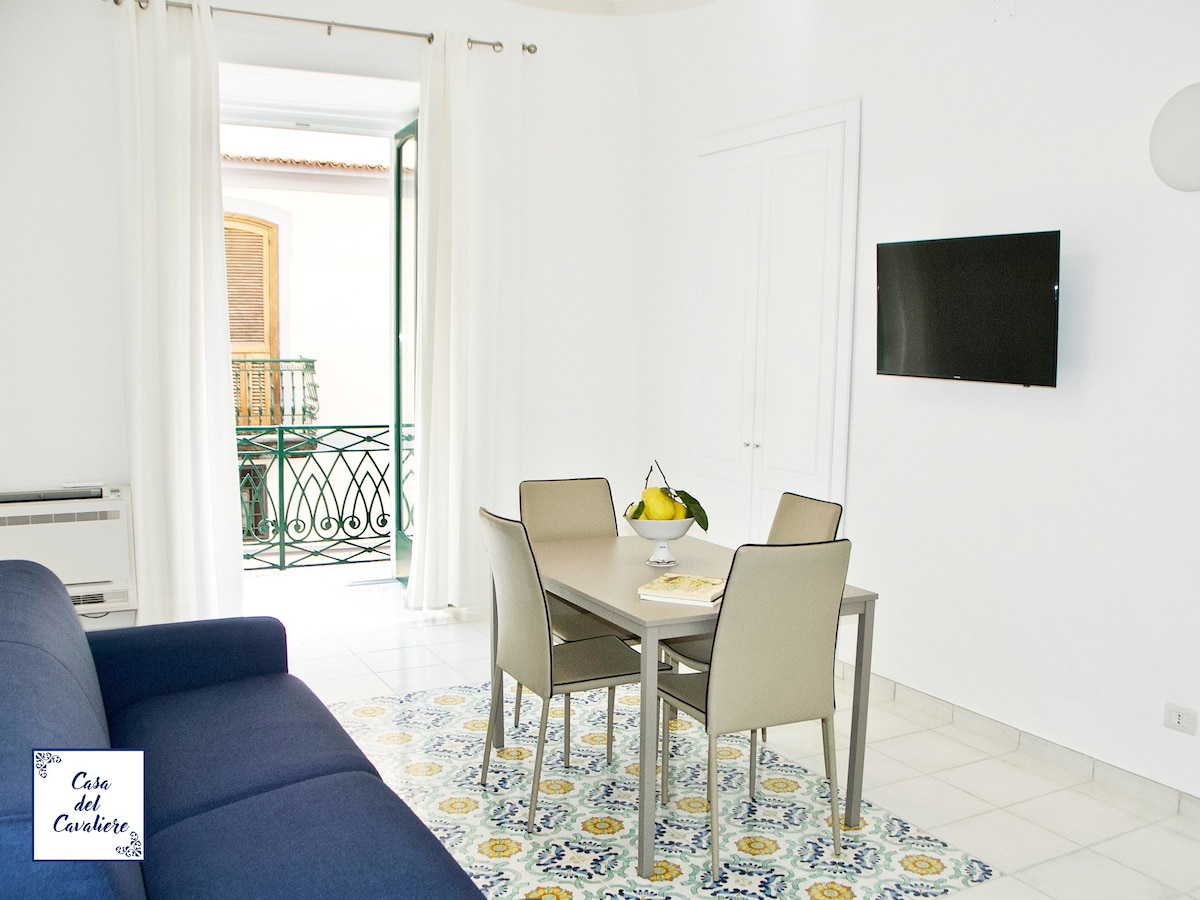 A bright living area is shown, featuring a blue sofa and a dining table with four chairs. A television is mounted on the wall, while large windows with white curtains offer views of a balcony. A colorful rug adds a decorative touch to the tiled floor.