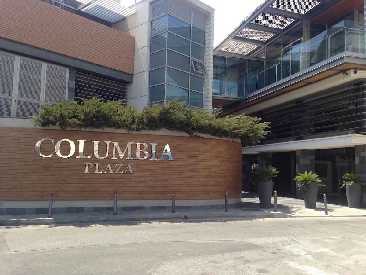 The exterior of Columbia Plaza is depicted, showcasing a modern building with large glass windows and a wood-paneled sign. Planters with greenery are positioned at the entrance, enhancing the contemporary design. The surrounding area appears spacious, with a paved pathway leading to the entrance.