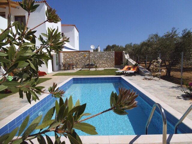 A private pool is surrounded by stone and tile, reflecting clear blue water under a sunny sky. Comfortable lounge chairs are positioned nearby, and olive trees line the background, providing a natural setting. The villa is visible, featuring white walls and a welcoming atmosphere.