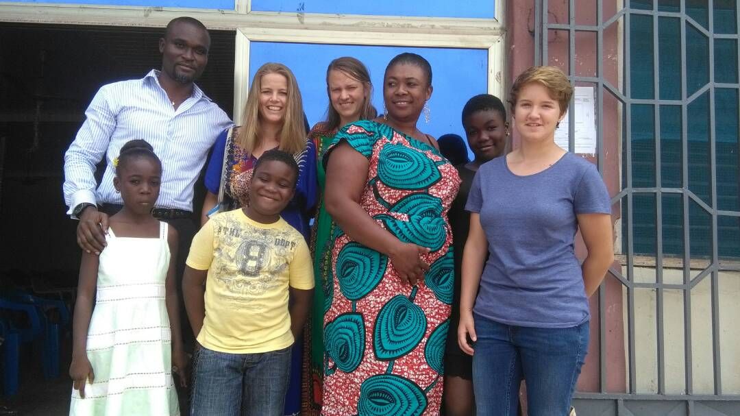 A group of five adults and children stands together outside a building with large blue windows. The individuals are dressed in colorful clothing, showcasing a variety of styles and patterns. A welcoming atmosphere is suggested by their friendly stance and smiles.