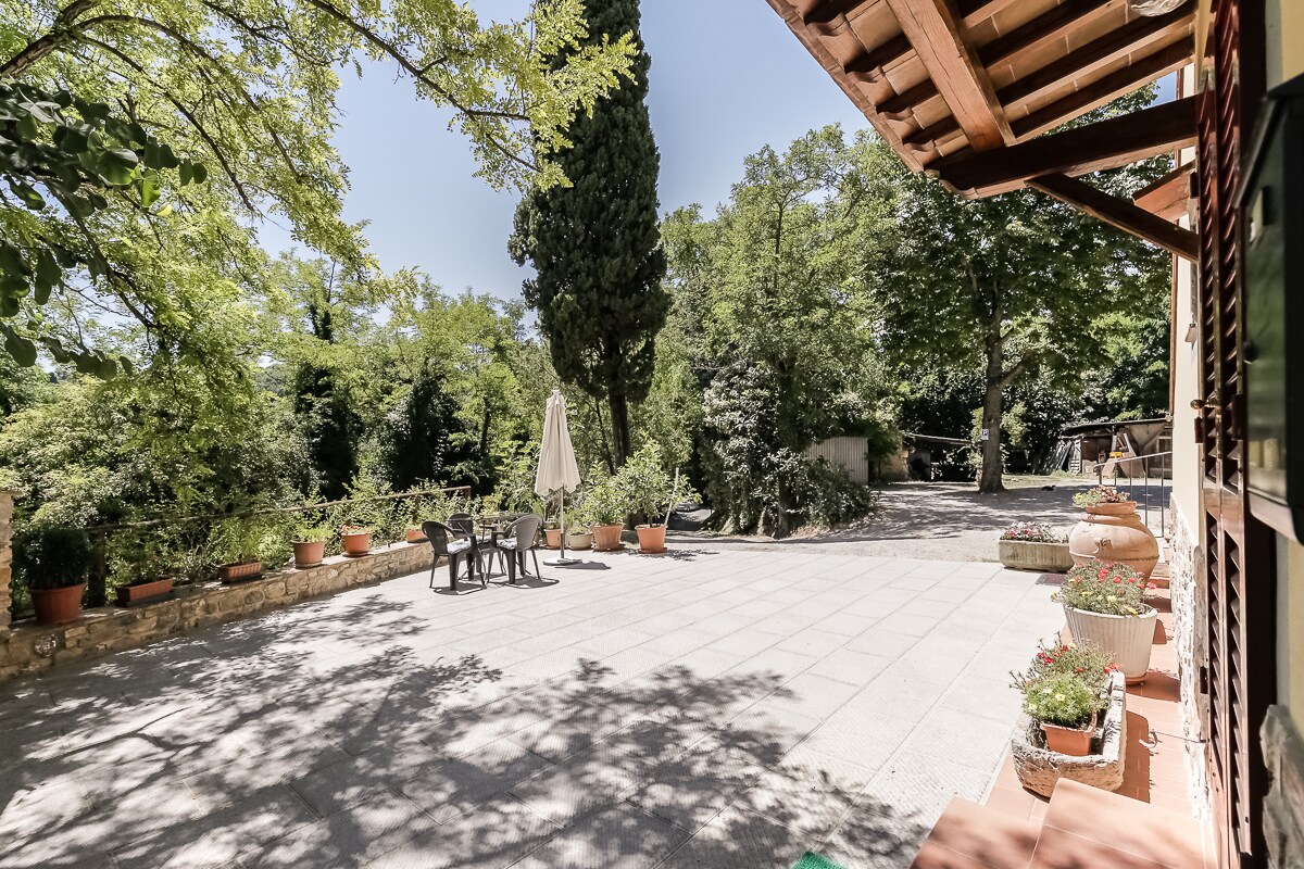 A spacious outdoor area features a stone-paved patio adorned with various potted plants. A table with chairs is positioned under a large umbrella, providing shaded seating. Lush greenery surrounds the area, enhancing the tranquil environment suitable for relaxation or dining outdoors.