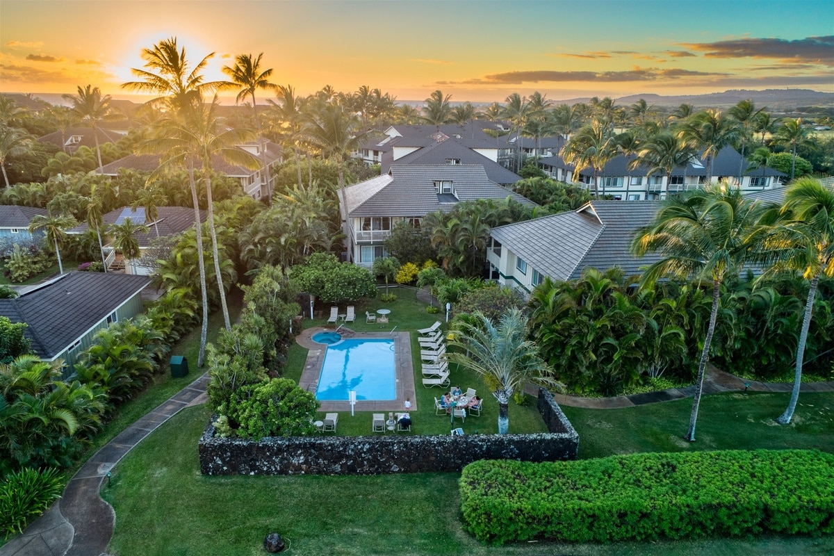 An aerial view showcases the beautiful grounds of a tropical resort. Lush palm trees surround a sparkling pool, where lounge chairs are arranged for relaxation. The sunset casts a warm glow over the landscape, enhancing the serene atmosphere of this inviting outdoor space.