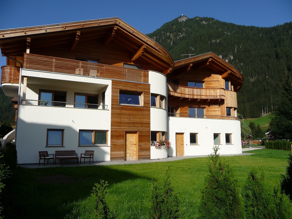 The exterior of the house showcases a blend of traditional alpine architecture and modern design. The building features wooden balconies and smooth white walls. Lush green grass surrounds the structure, complemented by a backdrop of a mountain landscape.