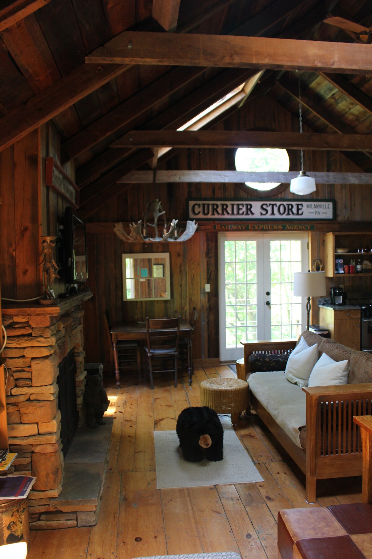 An inviting interior features rustic wooden beams and a stone fireplace. A cozy sofa is positioned facing the fireplace, supplemented by two wooden chairs and a small table. Natural light streams through glass doors leading to the outdoor area, enhancing the cabin's warm ambiance.