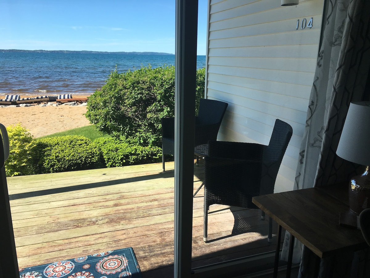 A view of the outdoor space is provided through sliding glass doors. A small deck features two wicker chairs facing the beach, with sandy shores and East Bay in the background. Lush greenery frames the view, enhancing the relaxing atmosphere.