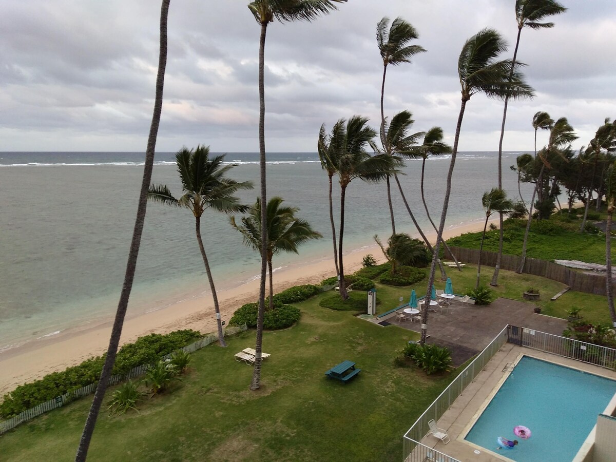 A shoreline view is presented, featuring a gentle beach with soft sand and a calm sea. Tall palm trees sway in the breeze, creating a tropical ambiance. A swimming pool and lounge area are visible on the property, surrounded by greenery and providing options for relaxation.