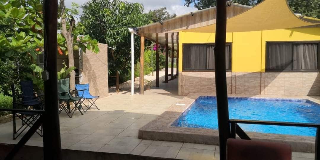 A private pool area features blue water surrounded by tiled decking. Two chairs are placed near the pool, with a shaded area provided by a large fabric canopy. A vibrant yellow structure is partially visible in the background, alongside lush greenery.