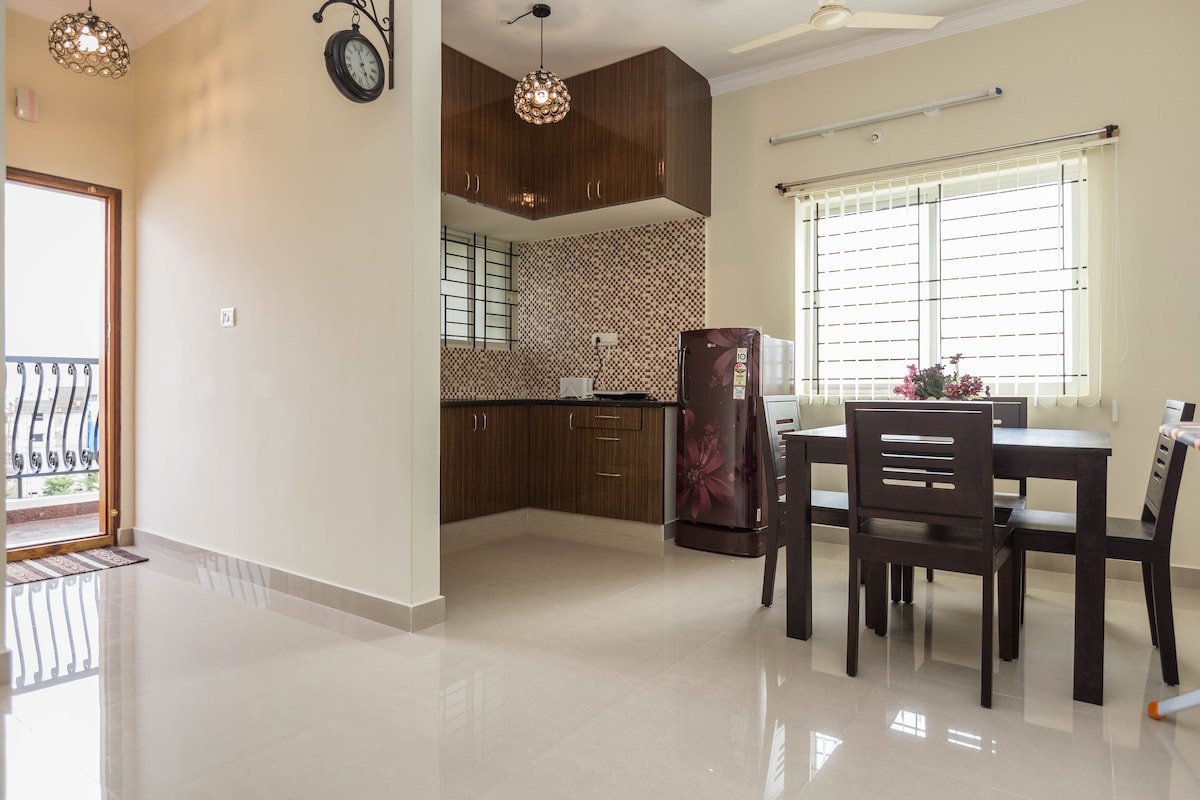 The bright kitchen area is highlighted by dark wood cabinetry and modern tile backsplash. A dining table with four chairs sits in the corner, adjacent to a refrigerator. Natural light enters through a window, enhancing the spacious and clean look of the room.