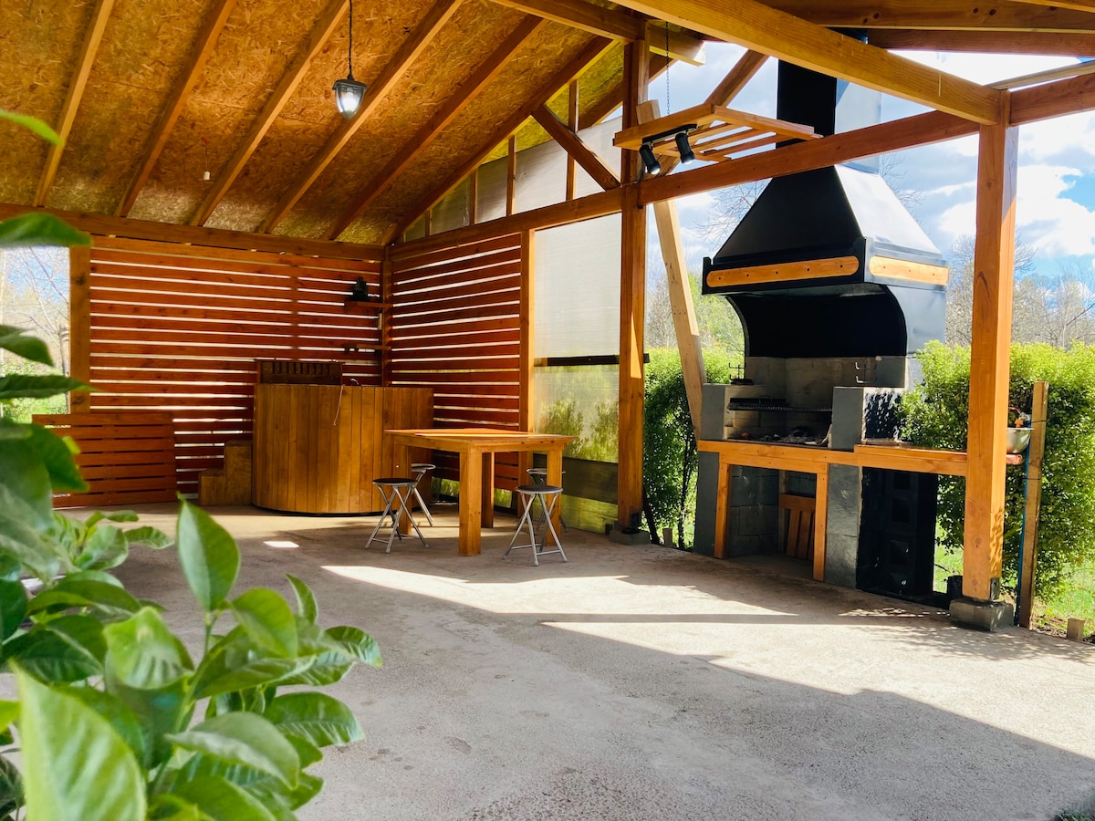An outdoor grilling area is presented with a wooden table surrounded by two folding chairs. A large barbecue is built into the structure, complemented by a traditional hot tub nearby. Natural greenery borders the space, and sunlight filters through the overhead beams.