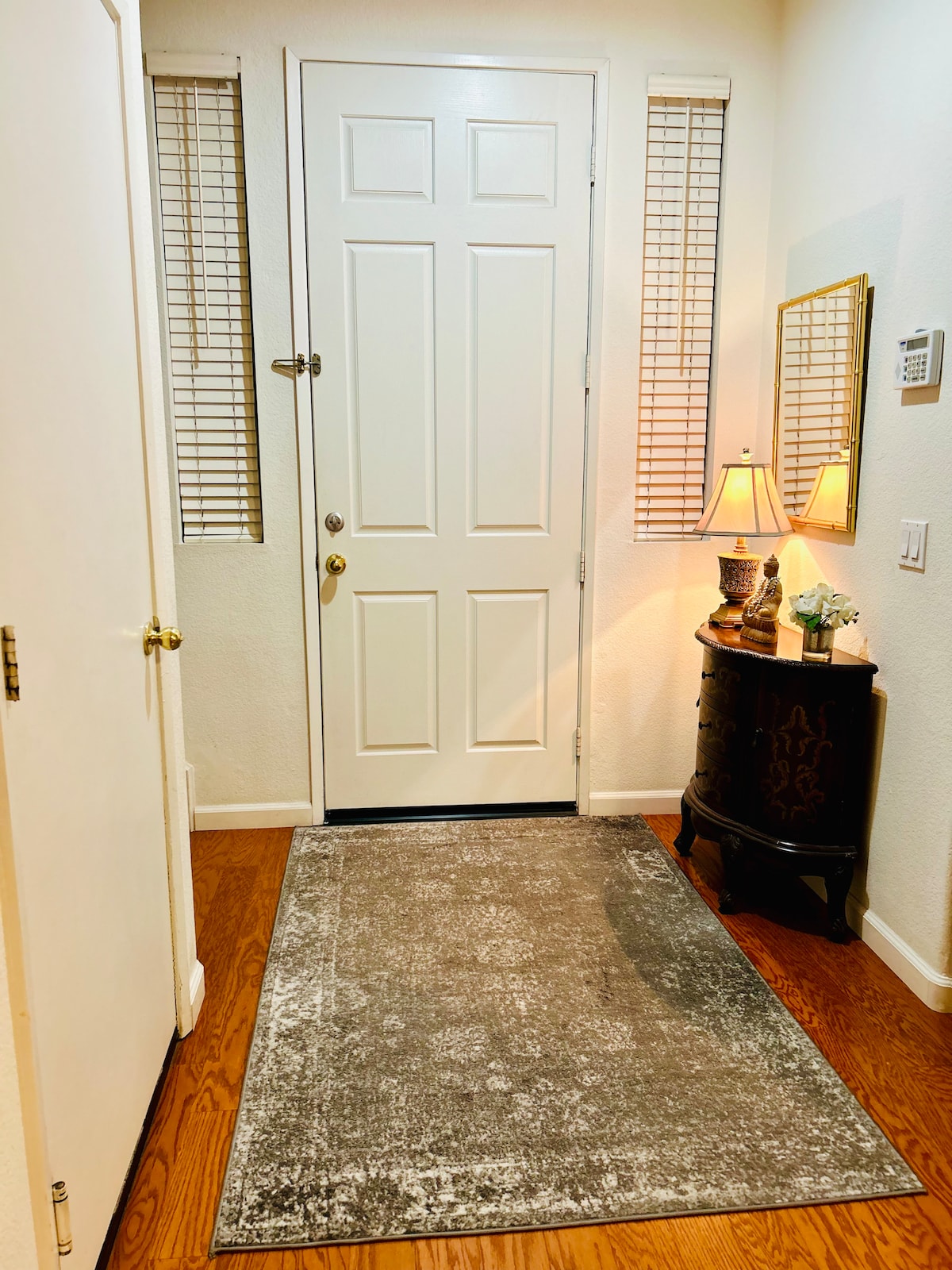 An entryway is presented featuring a white door with a glass panel handle. Two slender windows allow light to enter, and a small, dark wooden table is positioned beside the door, showcasing a lamp and decorative items. A textured area rug covers the wooden floor.