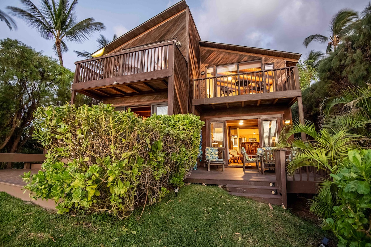 A two-story beach house features a wooden exterior surrounded by lush greenery. The upper balcony offers seating, while the lower level has large sliding glass doors leading into the living area. Palm trees are nearby, emphasizing the tropical setting.