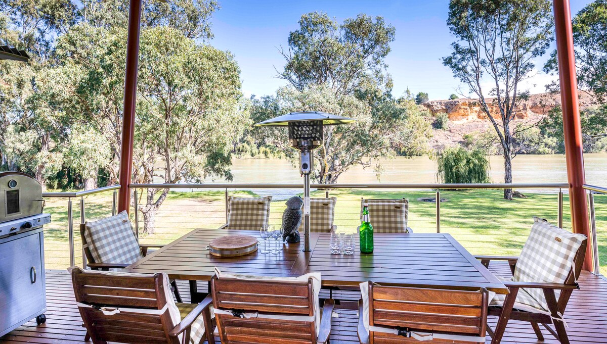 An outdoor dining area features a large wooden table surrounded by chairs, with views overlooking the river. A heater is positioned on the table, and a BBQ grill is visible in the background. Lush greenery frames the space, creating a serene atmosphere.