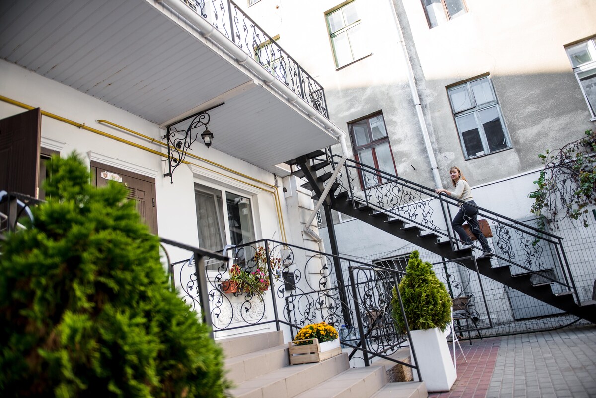 A set of elegant exterior stairs with decorative railings leads to a white-walled entrance of the building. Potted greenery and flowers add a touch of color to the courtyard, which is framed by surrounding buildings with multiple windows.