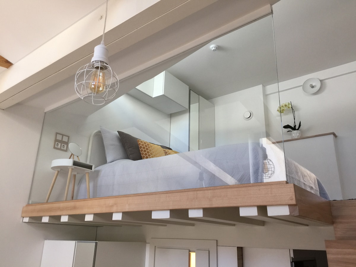 A lofted sleeping area is showcased above the living space, featuring a spacious bed with soft bedding. A wooden railing enhances safety while allowing visibility. A bedside table holds a lamp, and natural light enters through a nearby window, illuminating the room.