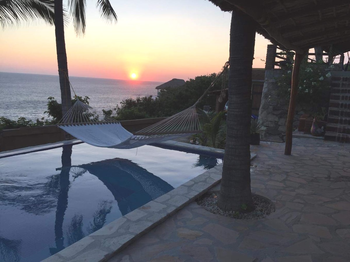 A tranquil view of the ocean during sunset is captured, with a hammock suspended near the edge of a serene pool. Palm trees gently sway in the breeze, and the warm colors of the sky reflect softly on the water's surface.