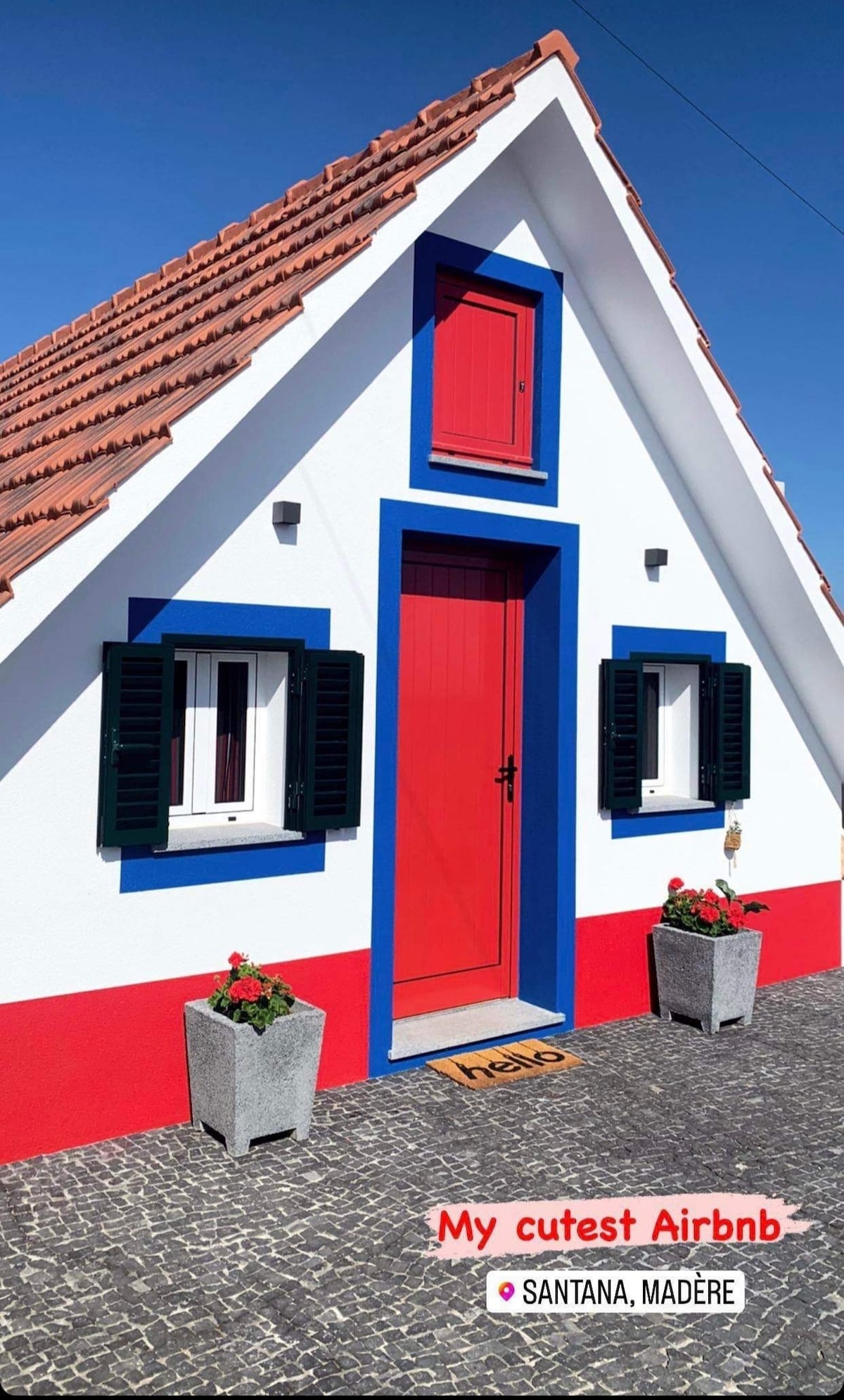 The charming exterior of the house features a red door complemented by blue trim and decorative shutters. Two flowers pots filled with vibrant plants are positioned at the entrance, set against a backdrop of white and red walls, characteristic of traditional Madeiran architecture.