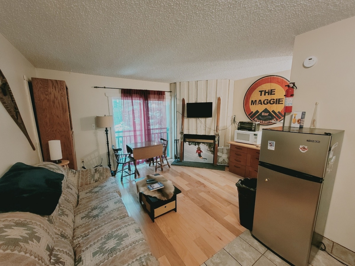 A cozy living space is shown, featuring a patterned couch and a wooden dining table with chairs. A flat-screen television is mounted above a fireplace. Natural light filters through a window with burgundy drapes, while a compact refrigerator and microwave provide kitchen essentials.